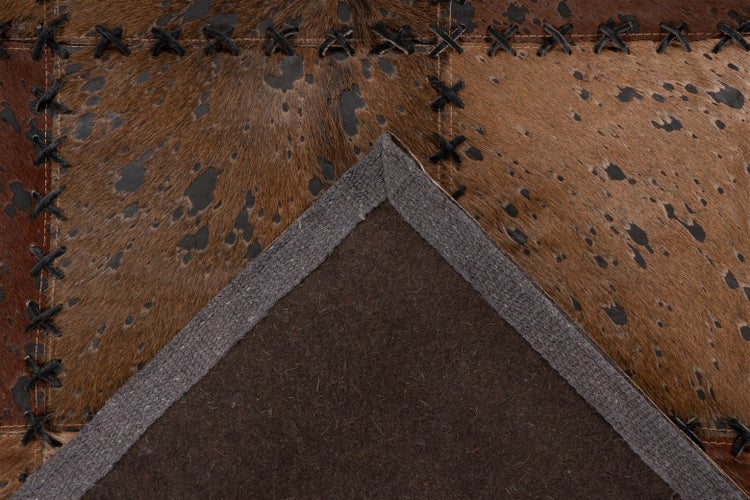 Detailansicht eines Patchwork-Teppichs aus Kuhfell mit Kreuzstich-Nähten und dunkler Filzrückseite mit grauem Rand.
