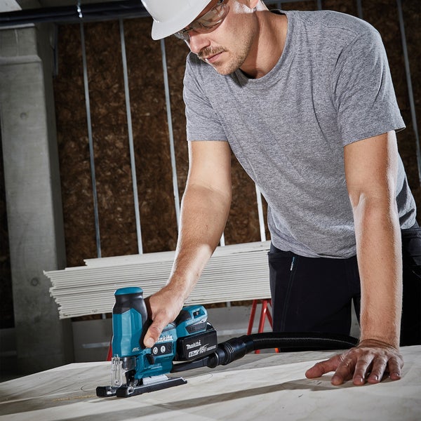 Ein Handwerker mit Schutzbrille und Helm sägt mit einer Makita Stichsäge ein Loch in eine Holzplatte.