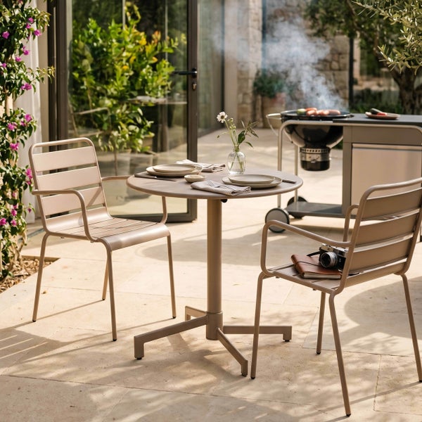 Modernes Bistro-Set mit rundem Tisch und zwei Metall-Armlehnenstühlen auf einer hellen Steinterrasse neben einem Grill und Pflanzen.