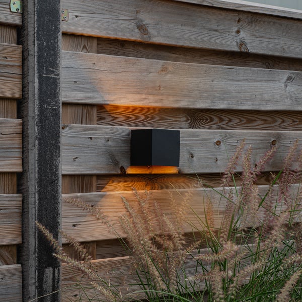 Quadratische, schwarze Außenwandleuchte an einem Holzzaun mit Lichtkegeln nach oben und unten sowie Gräsern im Vordergrund.