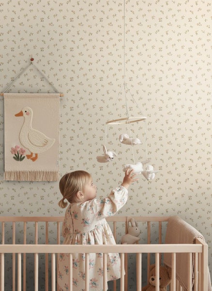 Helles Kinderzimmer mit Blumentapete, Holz-Kinderbett und Wandbehang mit Gans. Ein Kind greift nach einem Mobile mit Stofftieren.