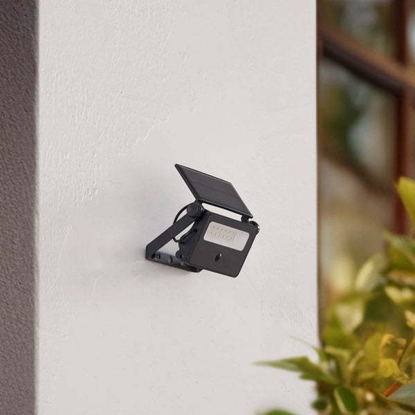 Schwarzer LED-Solarstrahler mit integriertem Solarpanel, montiert an einer weißen Außenwand.