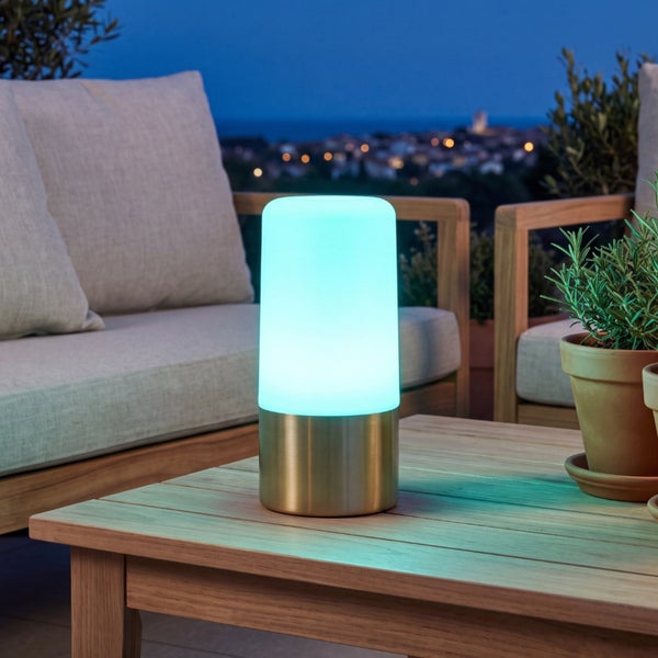 LED Tischleuchte für den Außenbereich mit Metallsockel und leuchtendem Zylinder auf einem Holztisch auf einer Terrasse in der Abenddämmerung.