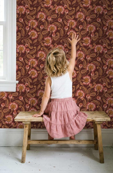 Ein Kind sitzt auf einer Holzbank vor einer Wand mit rot-rosa geblümter Mustertapete neben einem weißen Fensterrahmen.
