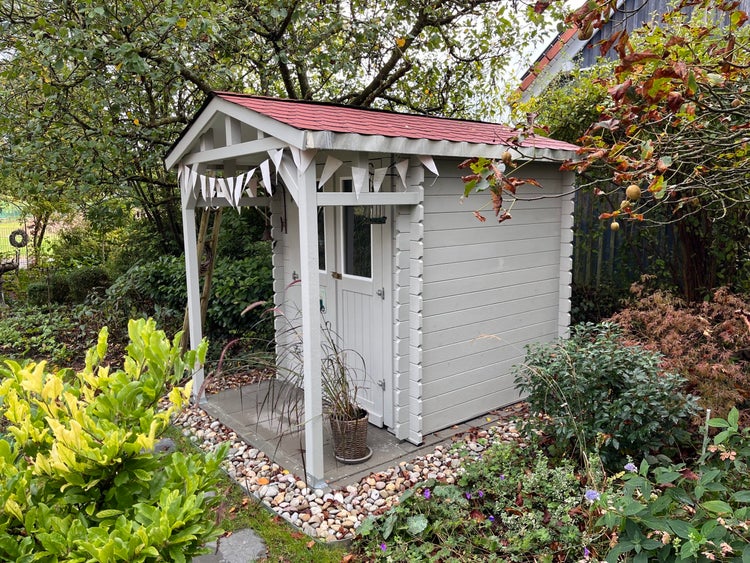 Gartenhaus aus Holz mit kleinem Vordach, rotem Schindeldach und weißer Fassade in einem üppig bepflanzten Garten mit Kieselsteinen.