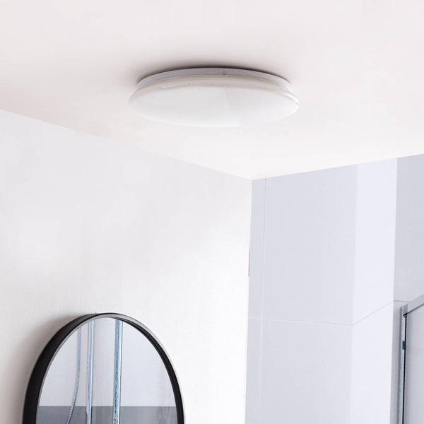 Runde weiße Deckenleuchte montiert an einer hellen Badezimmerdecke über einem runden Spiegel mit schwarzem Rahmen.