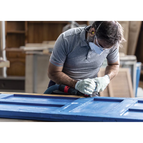 Ein Handwerker schleift mit einem Schleifgerät eine blaue Holztür. Er trägt dabei Schutzkleidung.