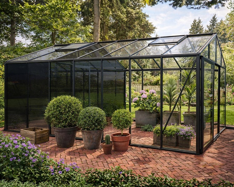 Großes, helles Gewächshaus aus Glas mit schwarzem Metallrahmen auf einer Backstein-Terrasse im Garten mit verschiedenen Topfpflanzen.