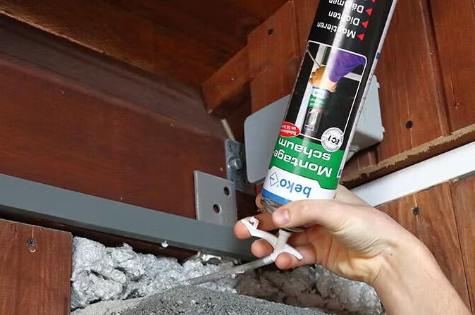 Hand trägt beko Montageschaum mit einem Adapterröhrchen in eine Fuge zwischen Holzgebälk und Mauerwerk auf.
