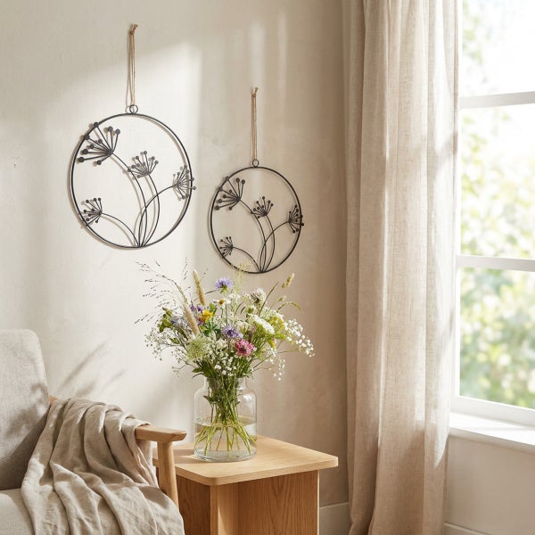 Zwei runde Wanddekorationen aus Metall mit Blumenmotiv hängen über einem Beistelltisch mit einer Glasvase voller Wildblumen in einem hellen Raum.