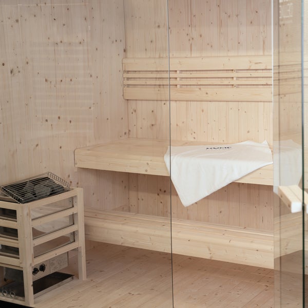 Innenansicht einer modernen Elementsauna mit Holzvertäfelung, Saunabänken, Saunaofen mit Steinen und einer Glastür.