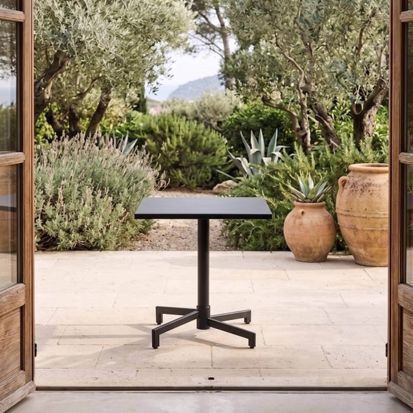 Quadratischer, schwarzer Bistrotisch mit Kreuzfuß auf einer sonnigen Steinterrasse, umgeben von mediterranen Pflanzen und Terrakottatöpfen.