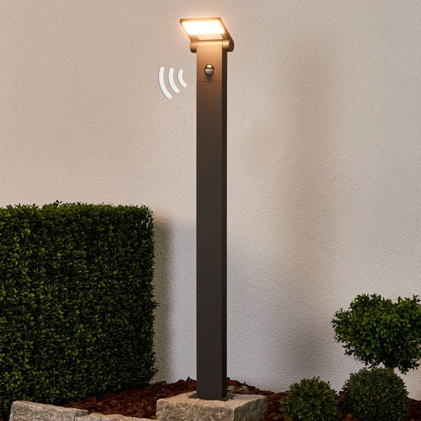 Moderne Außenstandleuchte mit Bewegungsmelder und LED-Panel, im Garten neben einer Hecke auf einem Stein montiert.