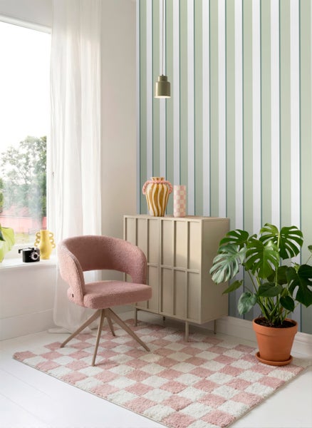 Hell eingerichtetes Zimmer mit vertikal gestreifter Vliestapete in Grün und Weiß, rosa Polstersessel, beigem Sideboard und einer Monstera-Pflanze.