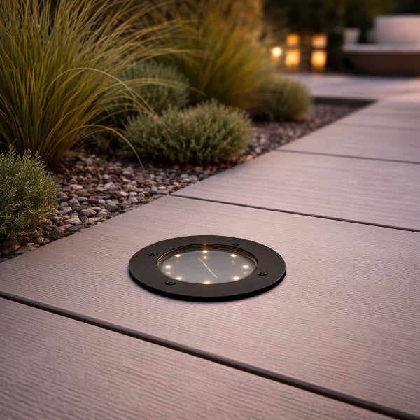 Solar-Bodeneinbaustrahler in einer Terrasse aus WPC-Dielen neben einem Gartenbeet mit Gräsern und Kieselsteinen.