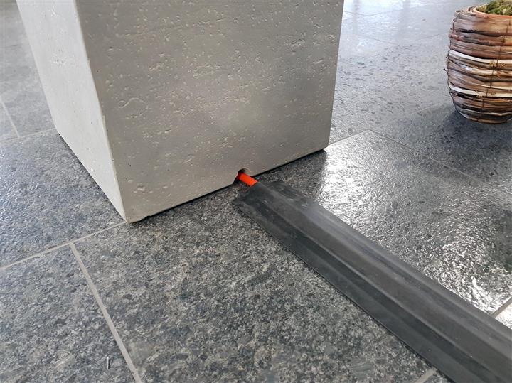 Kabelbrücke aus Gummi schützt ein Kabel auf einem Fliesenboden neben einem Pflanzkübel aus Beton.