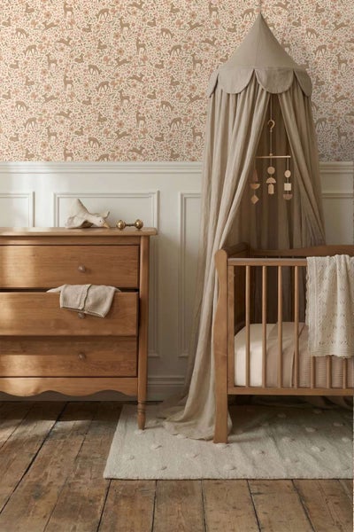 Ein gemütliches Babyzimmer mit Vliestapete mit Waldmotiven, einer Holzkommode, einem Kinderbett mit Betthimmel und einem strukturierten Teppich.