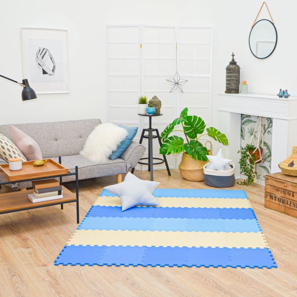 Heller Wohnraum mit blau-gelb gestreifter Schaumstoff-Puzzlematte auf Holzboden. Graues Sofa, Monstera in Korb und weiße Kaminumrandung sind sichtbar.