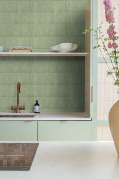 Moderne Kueche mit gruenen Wandfliesen im Hochformat, Holzregal, kupferfarbener Armatur und hellen Schrankfronten in organisierter Optik.