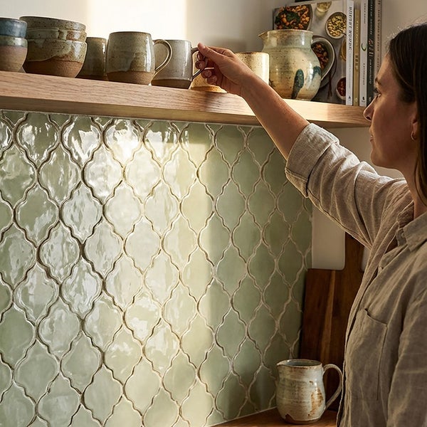 Eine Frau greift nach einer Tasse auf einem Holzregal vor einer Wand mit hellen, orientalischer Form gefliesten Küchenrückwand.
