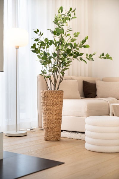 Künstliche Zimmerpflanze in einem hohen geflochtenen Übertopf aus Wasserhyazinthe in einem hellen Wohnzimmer neben einem Sofa.