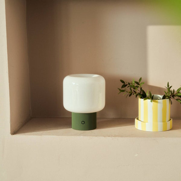 Moderne Tischleuchte mit abgerundetem weißem Schirm und grünem geriffeltem Sockel in einer Nische neben einer Pflanze.