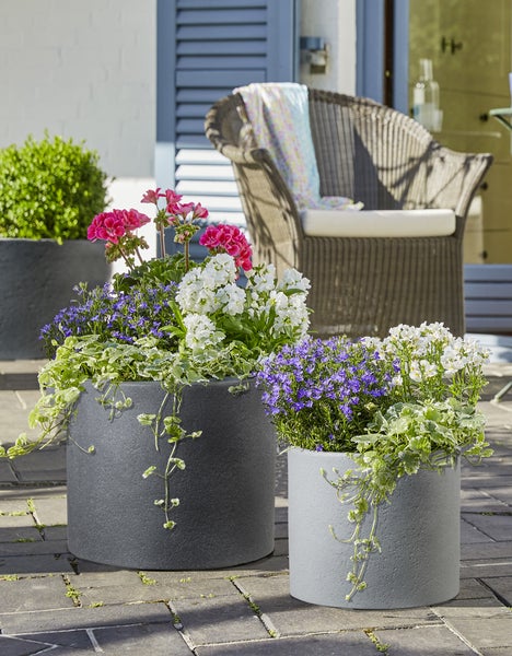 Zwei runde Pflanzkübel in verschiedenen Grautönen auf einer Terrasse mit Blumenbepflanzung und Gartenmöbeln im Hintergrund.