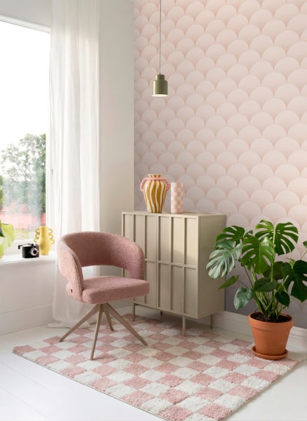 Helle Zimmereinrichtung mit Vliestapete im rosa Muscheldesign, rosa Sessel, beige Kommode und einem karierten Teppich.