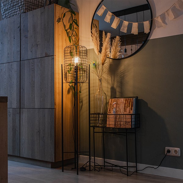 Einrichtungsbeispiel mit einer Stehlampe mit Drahtkorb, einem Beistelltisch, Dekoration und einem runden Spiegel.