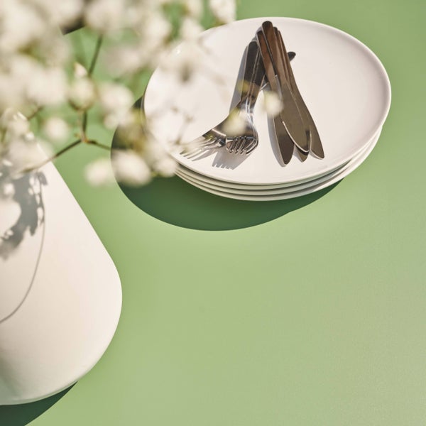 Stapel weißer Teller mit Besteck aus Edelstahl auf einem Tisch neben einer Vase mit weißen Blumen.