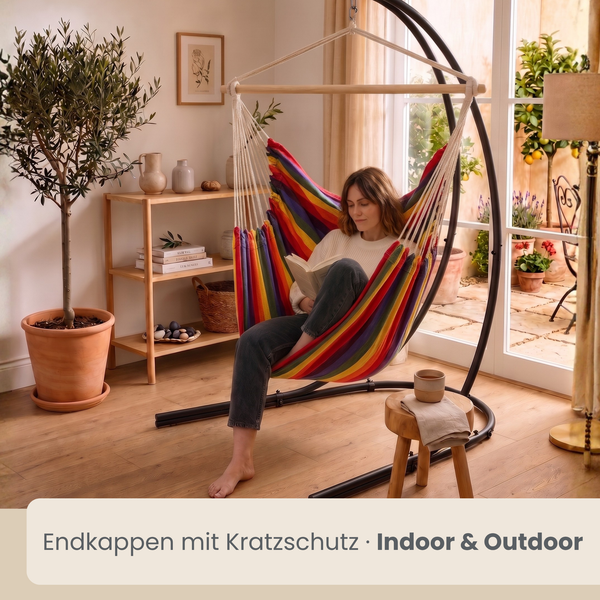 Frau sitzt in einem bunten Hängesessel mit Holzstreben an einem schwarzen Metallgestell in einem hellen Wohnraum mit Pflanzen.