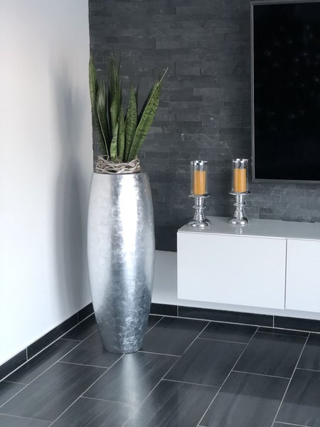 Modernes Wohnzimmer mit einer hohen silbernen Bodenvase mit Bogenhanf vor einer dunklen Steinwand und dunklen Bodenfliesen.