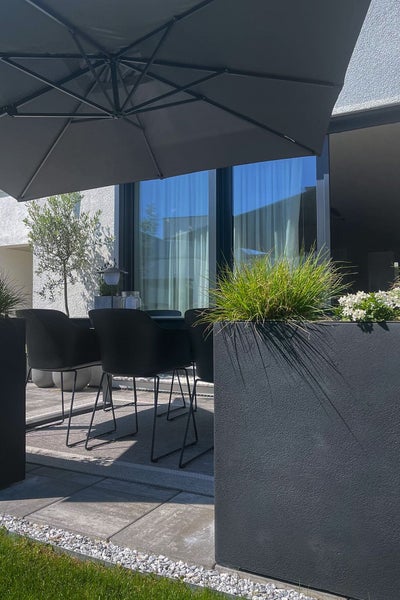 Moderne Terrasse mit großem Sonnenschirm, schwarzen Gartenstühlen und einem großen grauen Pflanzkübel mit Ziergras und weißen Blumen.