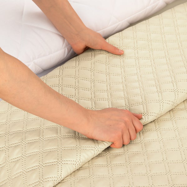 Eine Hand legt eine gesteppte Tagesdecke mit Rautenmuster auf ein weißes Kopfkissen.