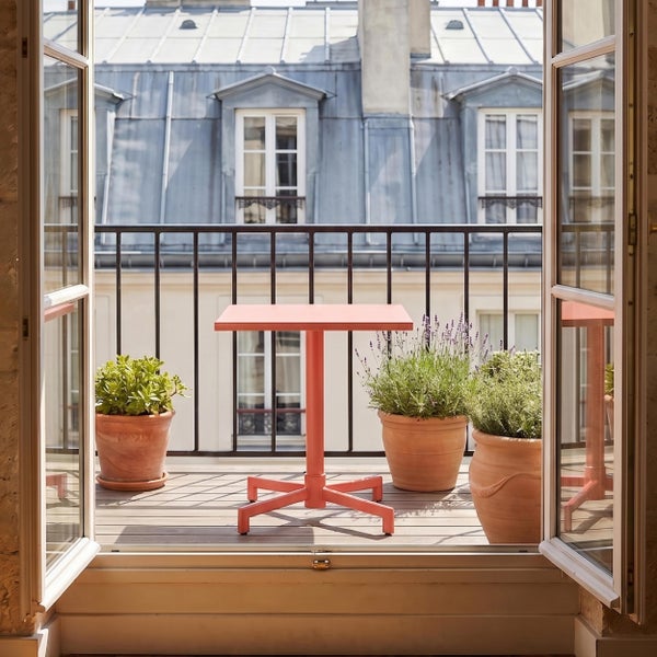 Kleiner Balkon mit einem quadratischen Bistrotisch, Tontöpfen mit Pflanzen und einem schwarzen Metallgeländer vor einem Stadtgebäude.