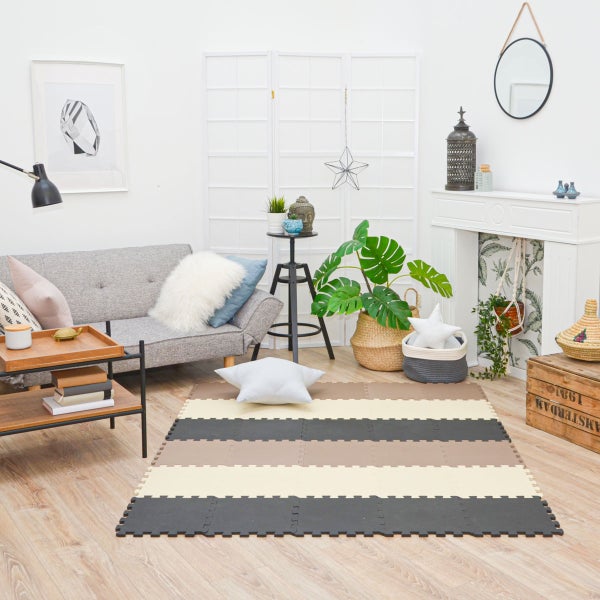Helles Wohnzimmer mit grauem Sofa, einer Monstera-Pflanze und einer gestreiften Schaumstoff-Bodenschutzmatte in der Raummitte.