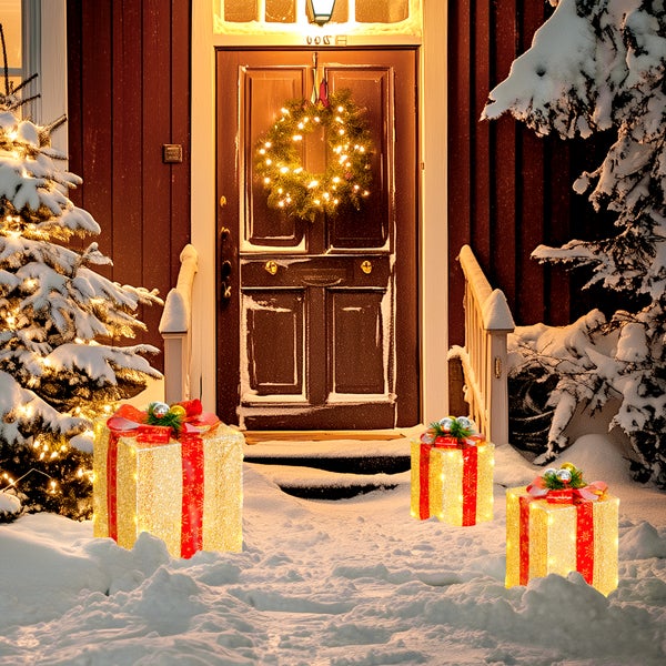 Drei beleuchtete Weihnachtsgeschenk-Dekorationen in einem verschneiten Garten vor einer Haustür mit Kranz und schneebedeckten Tannen.