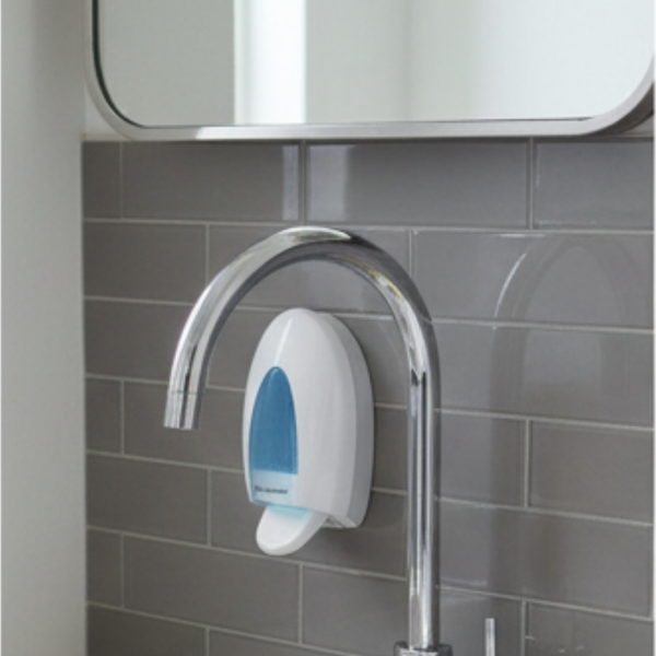 Wandmontierter Seifenspender weiß mit blauem Sichtfenster an grauer Fliesenwand hinter verchromter Armatur.