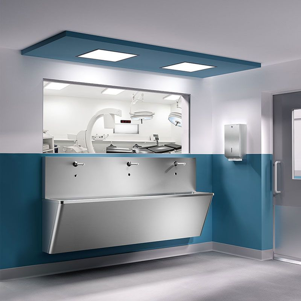 Chirurgisches Waschbecken aus Edelstahl mit drei Armaturen in einem hellen klinischen Raum vor einem Fenster zum Operationssaal.