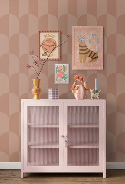 Rosa Metallvitrine mit Riffelglas-Türen vor geometrischer Bogentapete. Dekorative Vasen und eingerahmte Bilder sorgen für ein helles, ordentliches Bild.