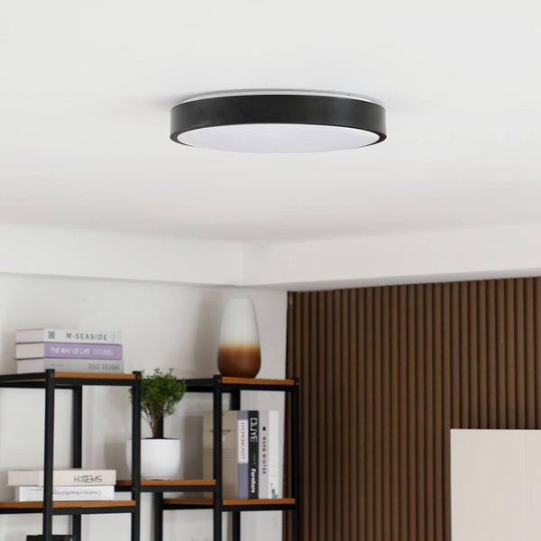 Moderne runde LED-Deckenleuchte mit schwarzem Rahmen und weißem Diffusor in einem hellen Raum mit Regal und Holzpaneelwand.