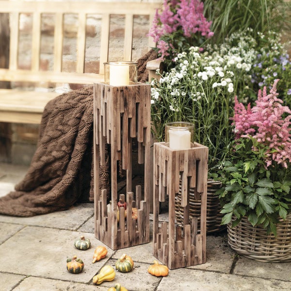 Zwei hohe Windlichter aus Holz in moderner Stäbchenoptik mit Kerzenglas für den Garten, dekoriert mit kleinen Kürbissen auf einer Steinterrasse.