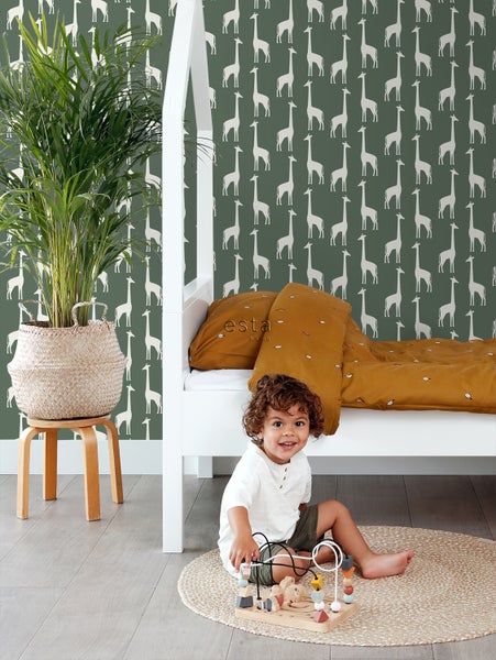 Kinderzimmer mit weißem Bett, Zimmerpflanze und einem Kind, das mit einem Spielzeug spielt, vor einer grünen Tapete mit Giraffenmuster.