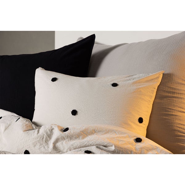 Weißes Kissen mit schwarzen Noppen aus Baumwolle als Teil eines Bettwäsche-Ensembles in einem hellen Schlafzimmer.