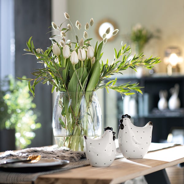 Dekorative Szene mit einem Blumenstrauß in einer Glasvase und zwei Keramikfiguren in Hühnchenform auf einem Tisch