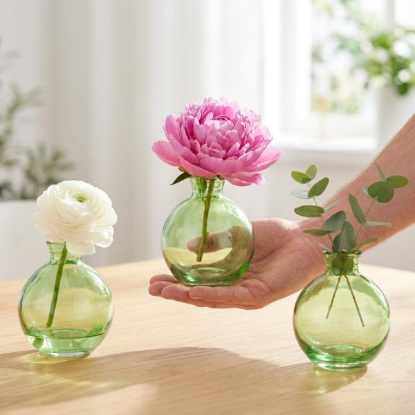 Drei kleine, bauchige Glasvasen auf einem Holztisch, eine davon wird von einer Hand gehalten, dekoriert mit Blumen und Eukalyptuszweigen.