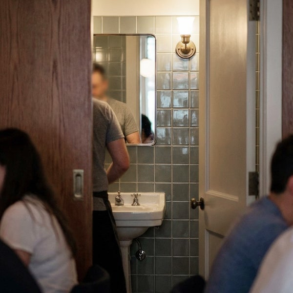 Ein Badezimmer mit einem Waschbecken an einer gekachelten Wand, einem Wandspiegel und einer Wandleuchte, gesehen durch eine halb geöffnete Tür.