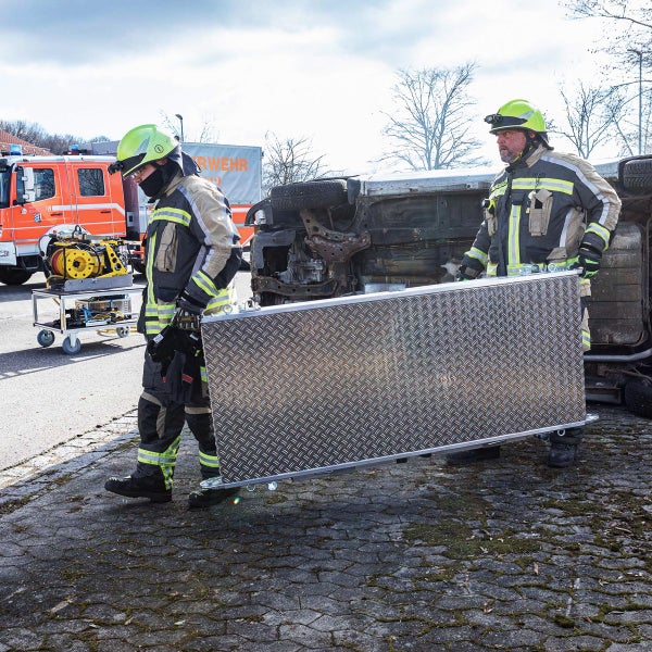 Zwei Feuerwehrleute tragen eine Metallplatte vor einem umgestürzten Auto.