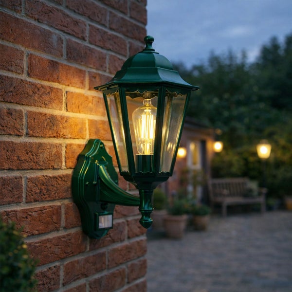 Klassische grüne Außenwandleuchte in Laternenform mit Bewegungsmelder und warmem LED-Licht, montiert an einer Ziegelwand.