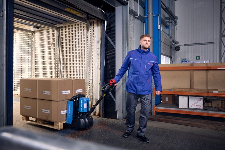 Ein Mann in blauer Arbeitsjacke zieht mit einem elektrischen Hubwagen eine Palette mit Kartons aus einem Container in einem hellen Lager.